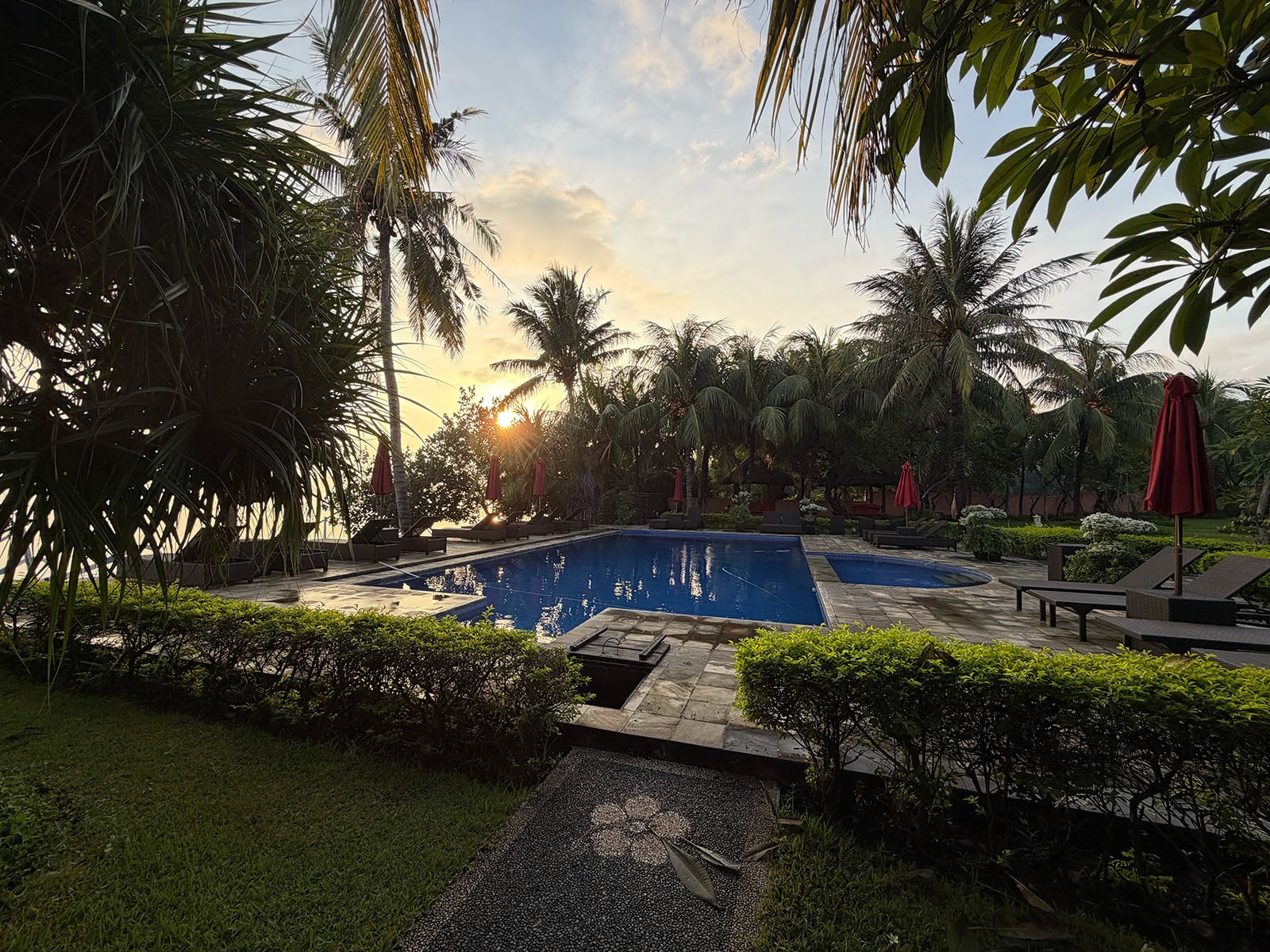 A pool lit from within at dusk, lawn and coconut palms around it, the sky still orange behind the trees