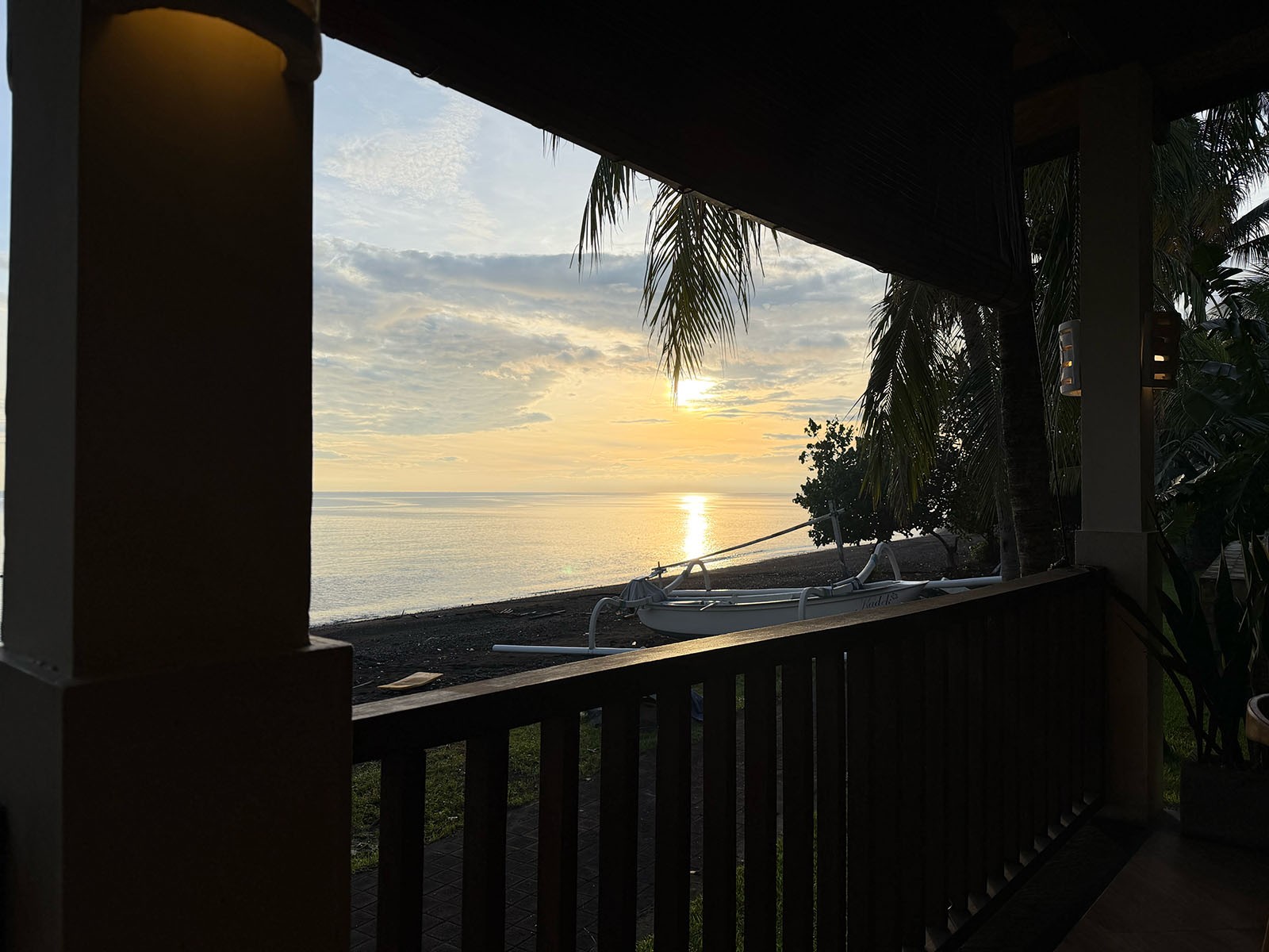 The sea seen from a shaded balcony at sunset, a dark timber rail across the foreground