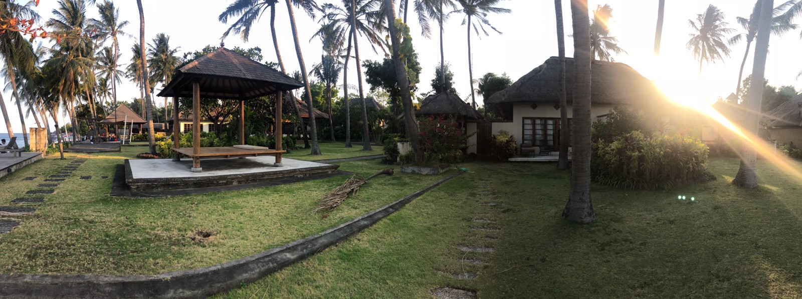 Low sun through palms across a lawn, an open pavilion and thatched cottages on the far side