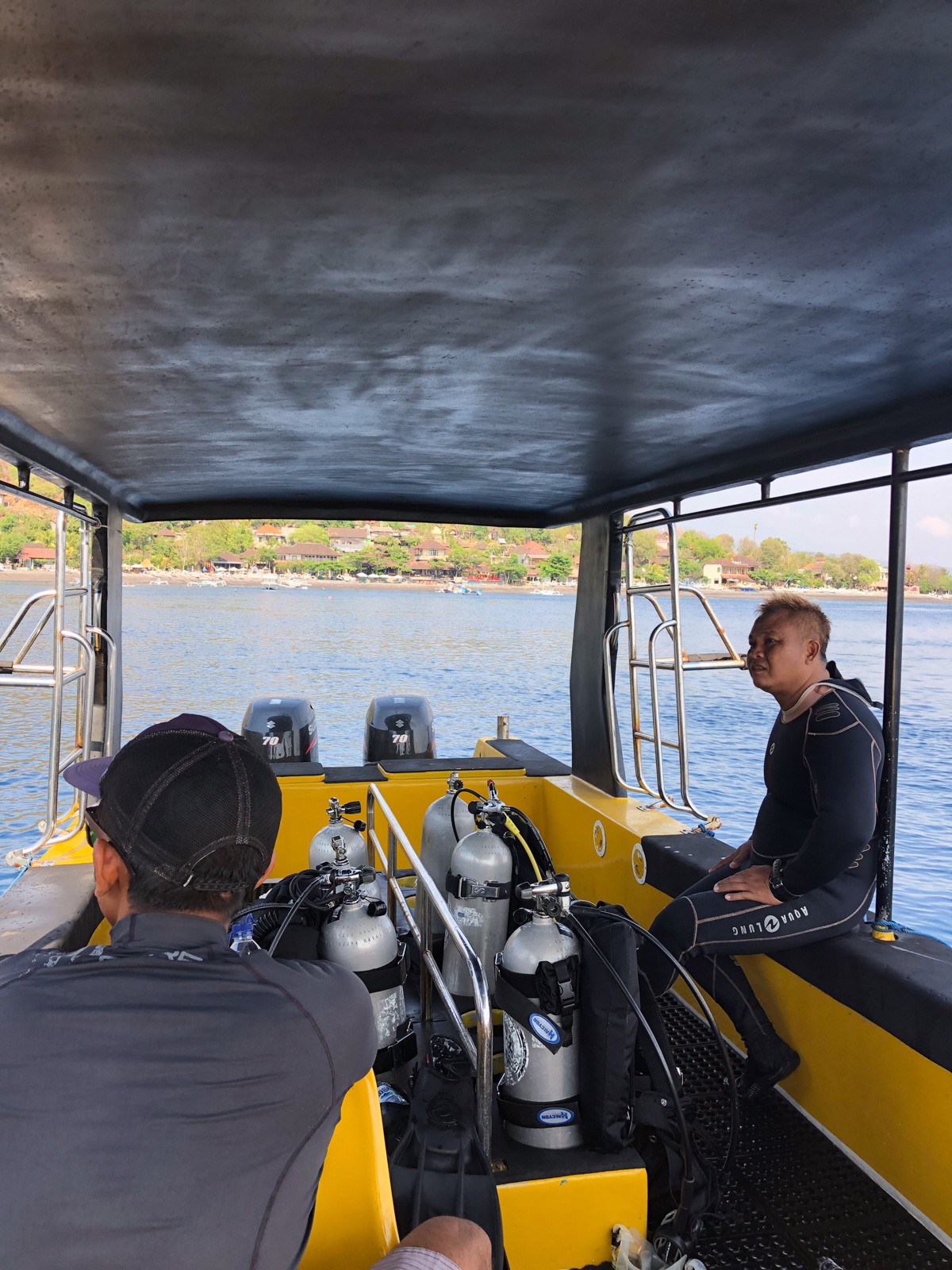 The covered deck of a dive boat, aluminium tanks racked along the yellow gunwale, a crew member sitting at the stern with the Bali coast behind