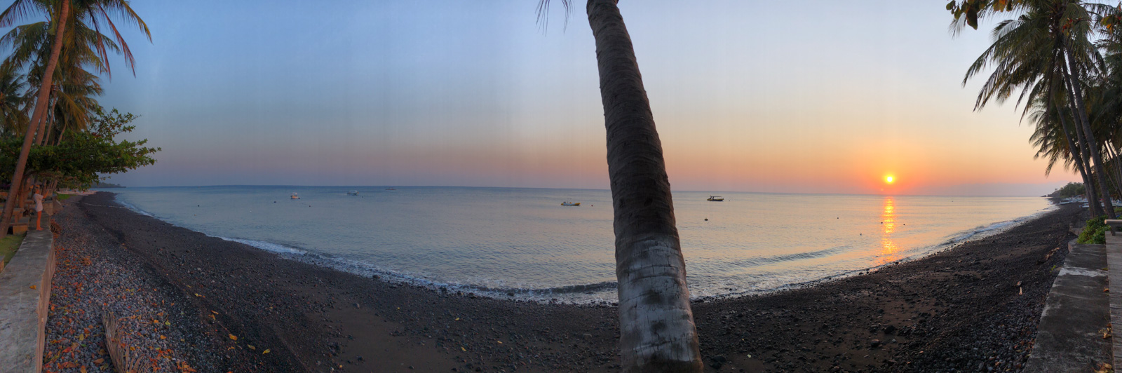 The black volcanic beach at first light, boats lying off and the sun coming up between the palms.