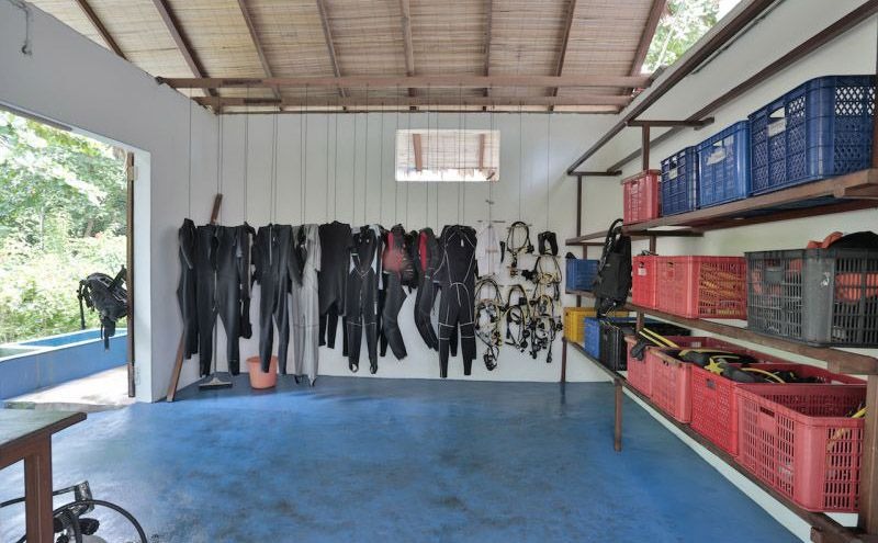 A gear room with wetsuits hung in a row along one wall, stacked crates and a workbench opposite