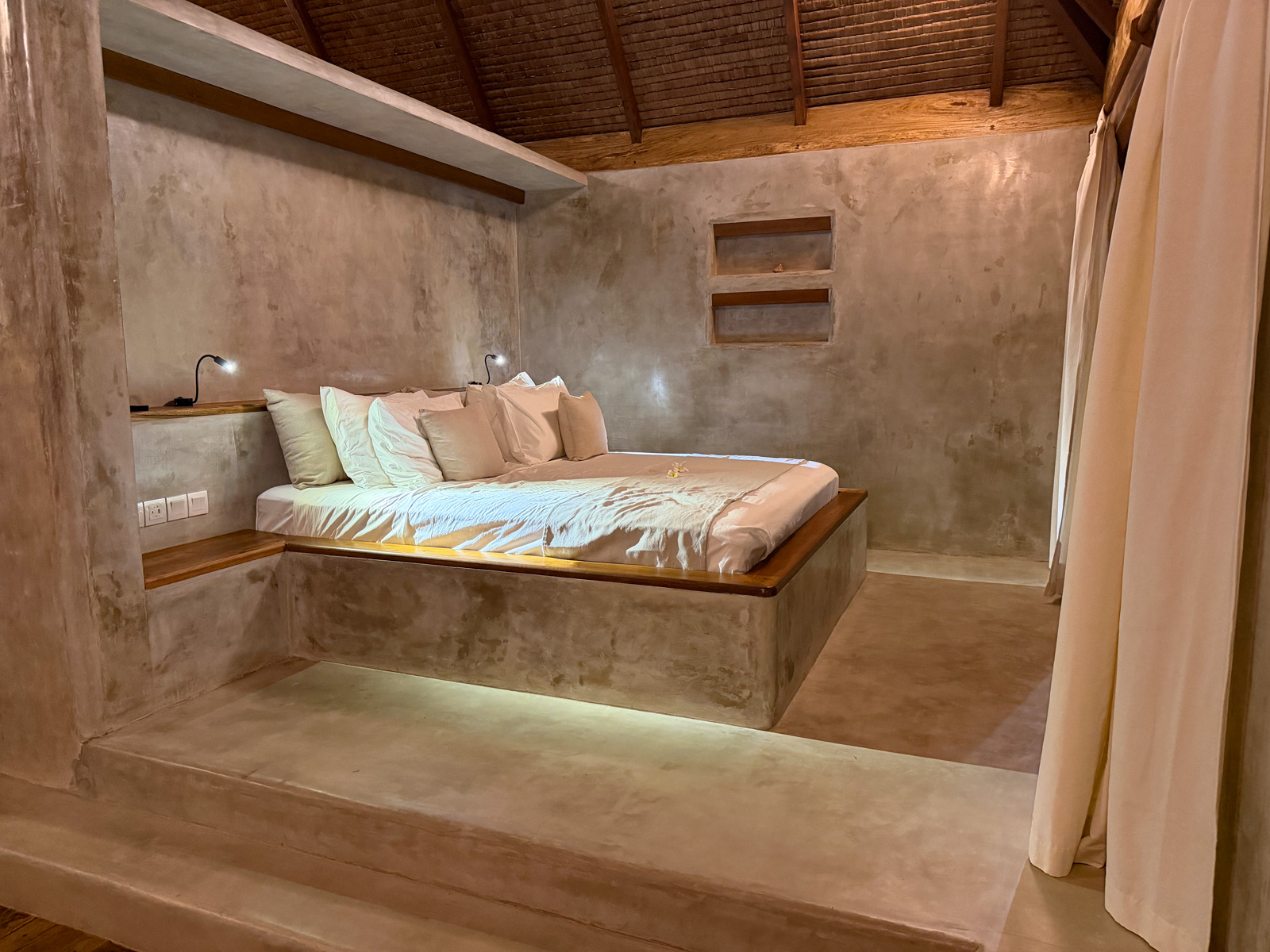 A bedroom in polished concrete, a low platform bed under a shelf recess and a muslin curtain drawn back.