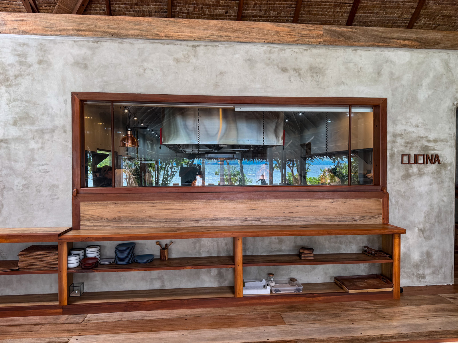 The open kitchen seen through a wide serving window, CUCINA lettered on the wall and a long timber counter beneath.