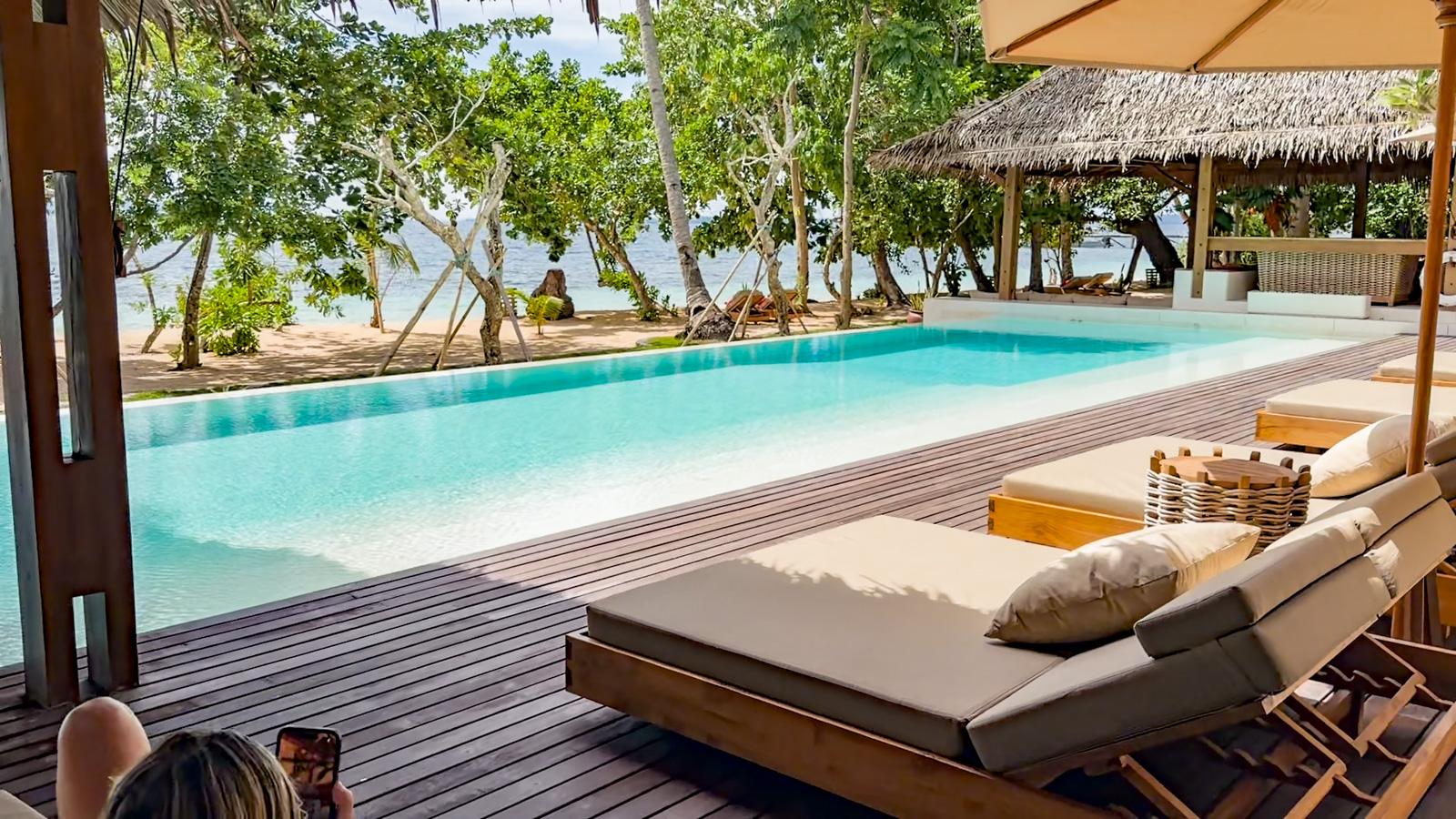 A long pool on a timber deck, loungers under a thatched pavilion and the sea through the palms behind.