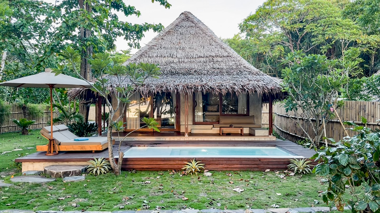 A thatched villa on a lawn with its own plunge pool and a parasol beside it.