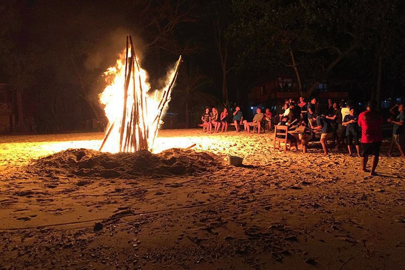 A driftwood bonfire burning on a beach at night, a ring of people watching from the sand