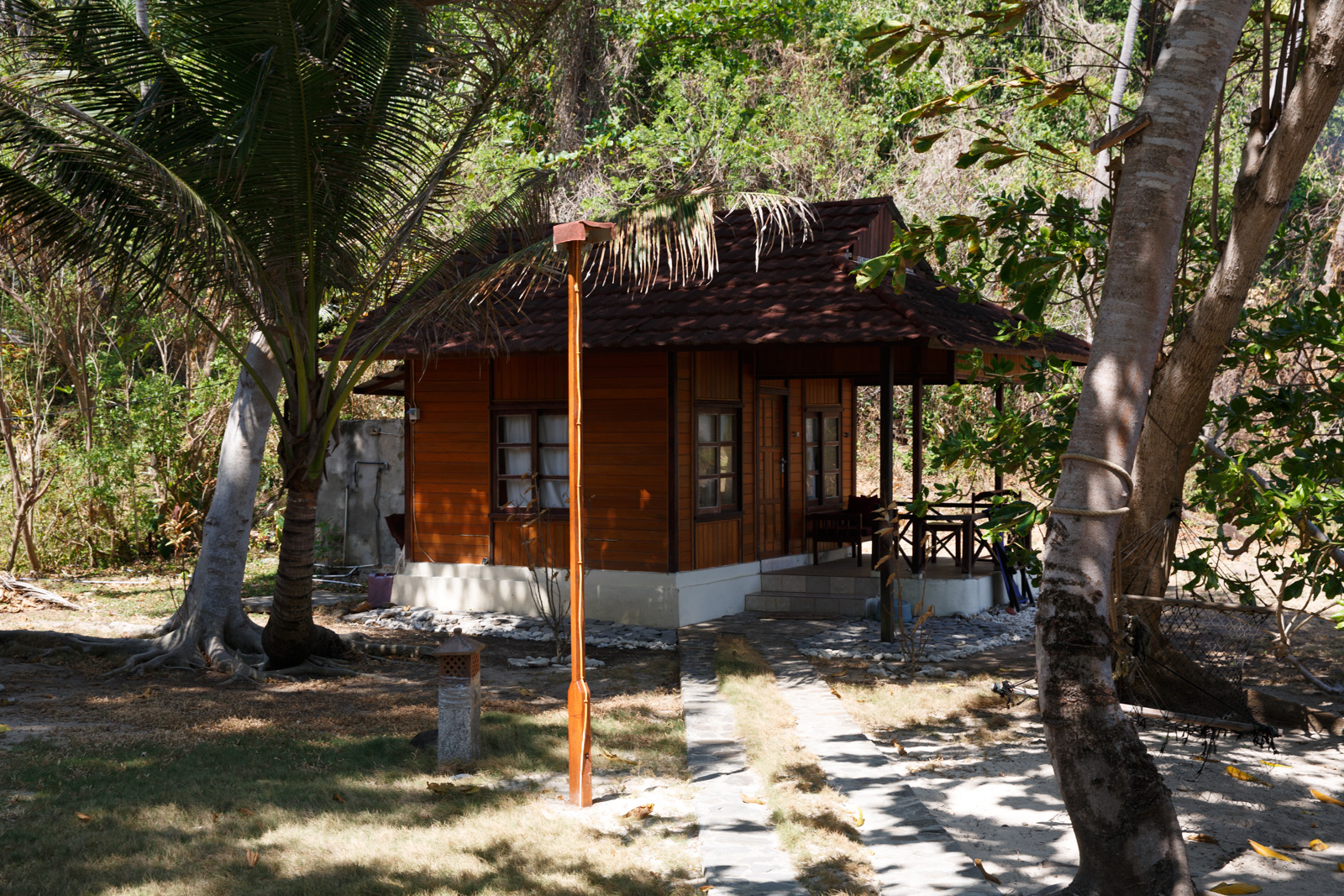 A timber cottage under a thatched roof standing in the shade of the palms, its verandah furnished and looking onto the sand.