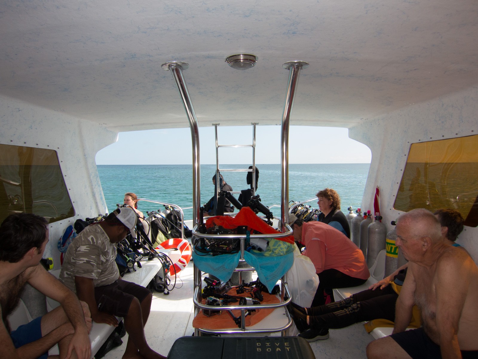 The inside of a covered dive boat, divers in wetsuits sitting along both benches with gear between them