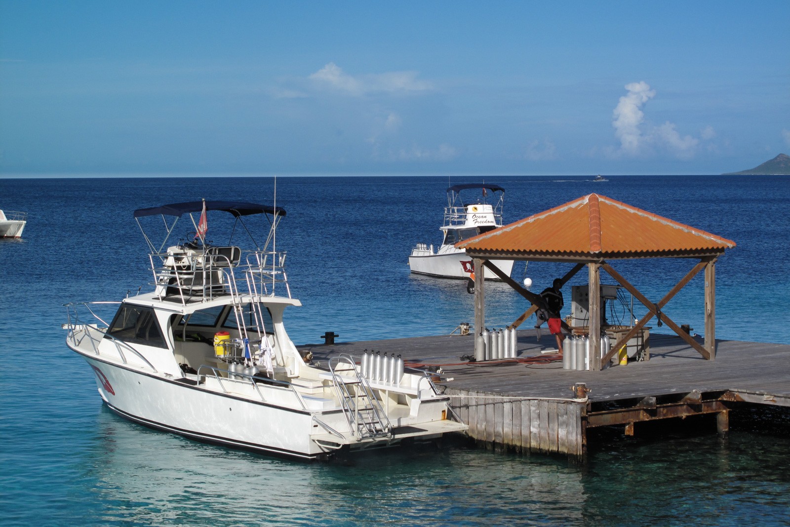 A white dive boat tied alongside a wooden dock with a thatched shade hut, flat blue water and low hills behind