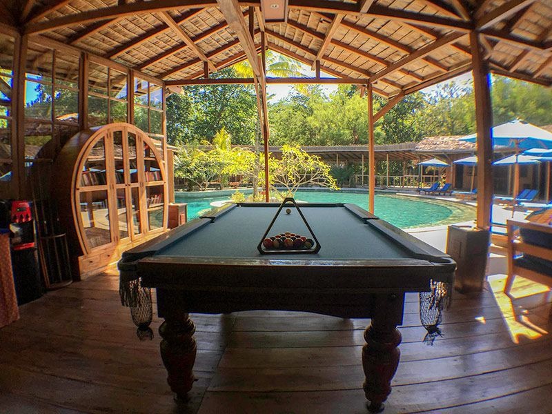 An open-sided timber pavilion with a pool table, the swimming pool and blue parasols beyond it