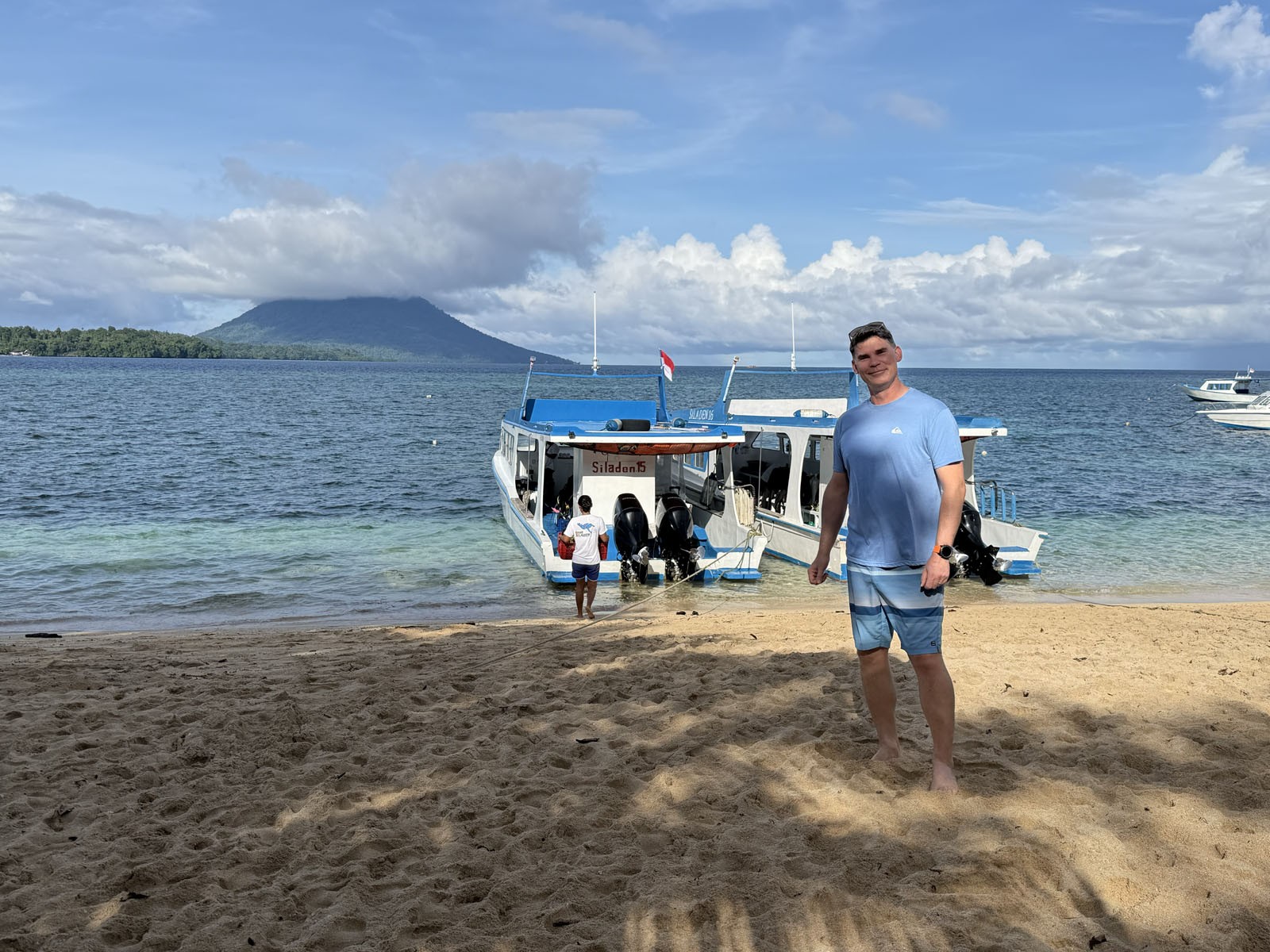 A man standing on the sand beside a white dive boat pulled up in the shallows, the volcanic cone of Manado Tua across the water
