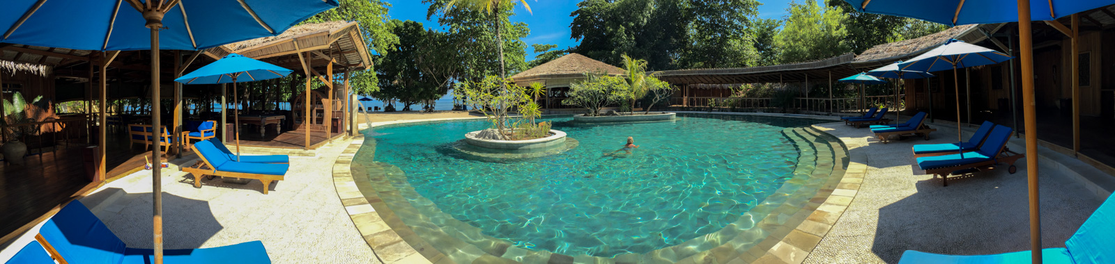 The pool in the middle of the day, blue parasols and loungers round three sides and one swimmer crossing it.