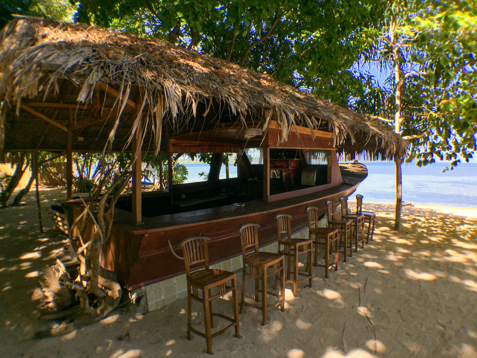 A beach bar built into a boat hull under a thatched roof, stools drawn up along it in the shade.