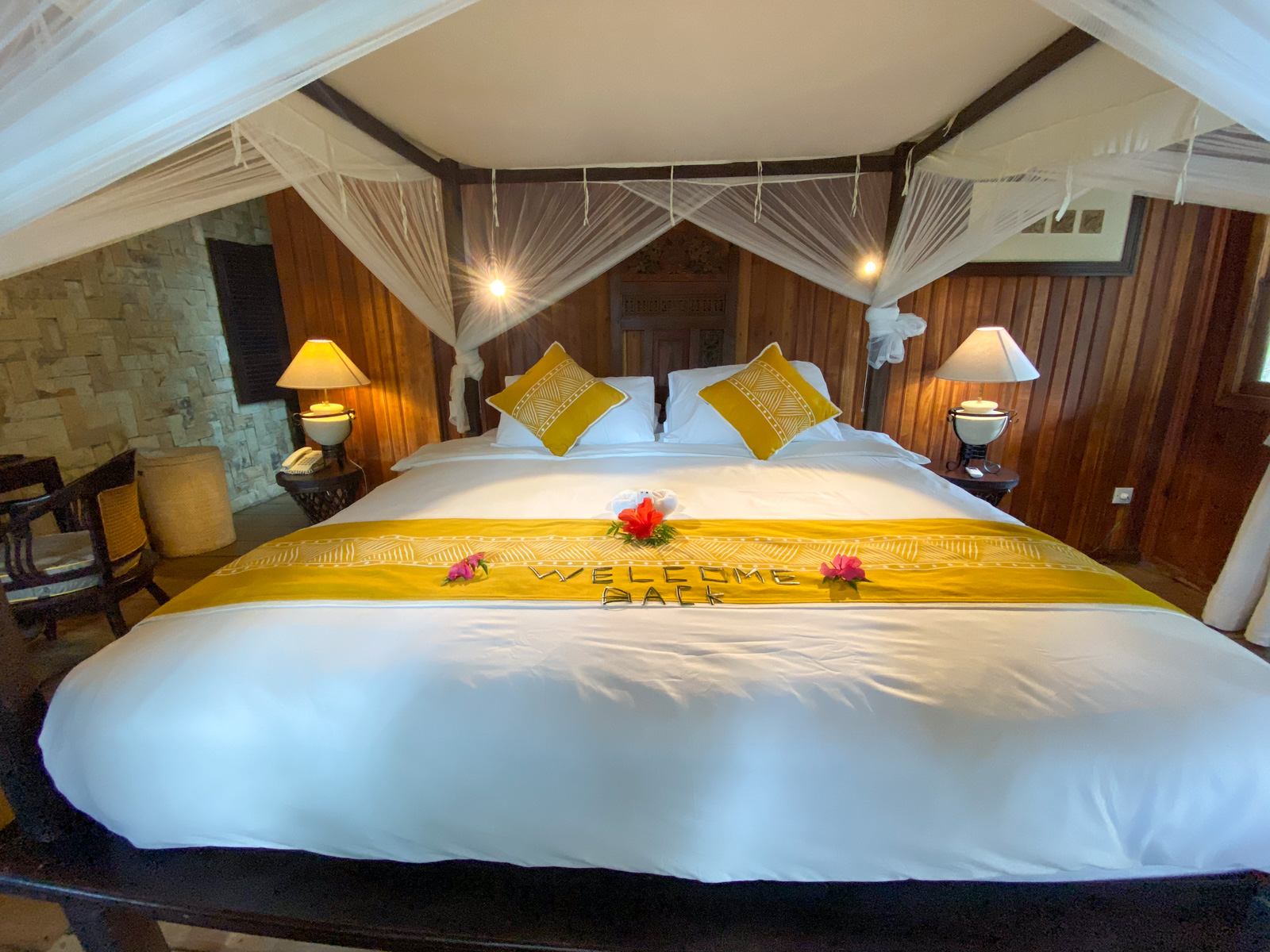 A four-poster bed under a mosquito net, a welcome runner and flowers laid across the white cover.