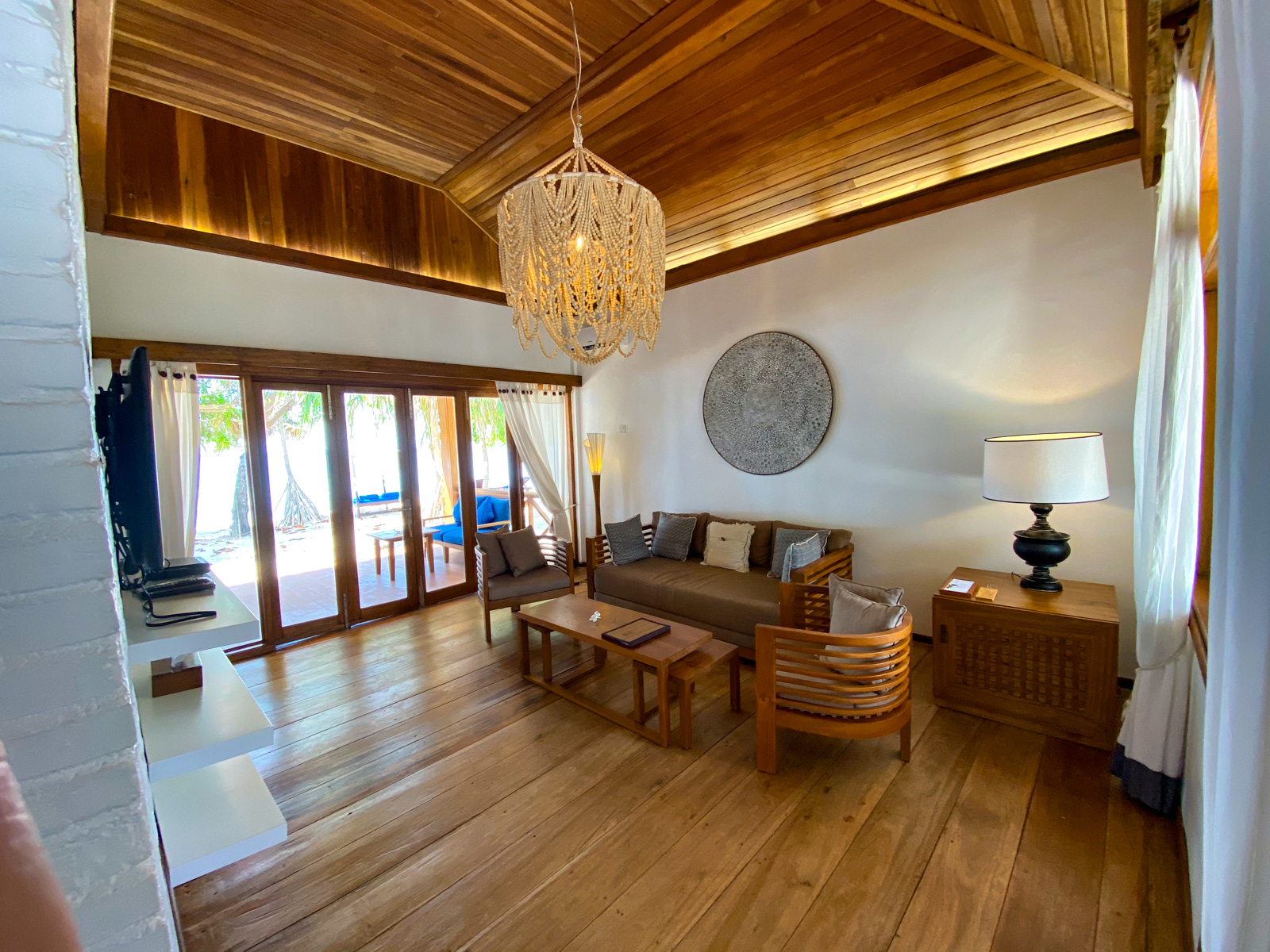 A sitting room under a high timber ceiling, a shell chandelier over the sofa and doors open to the terrace.