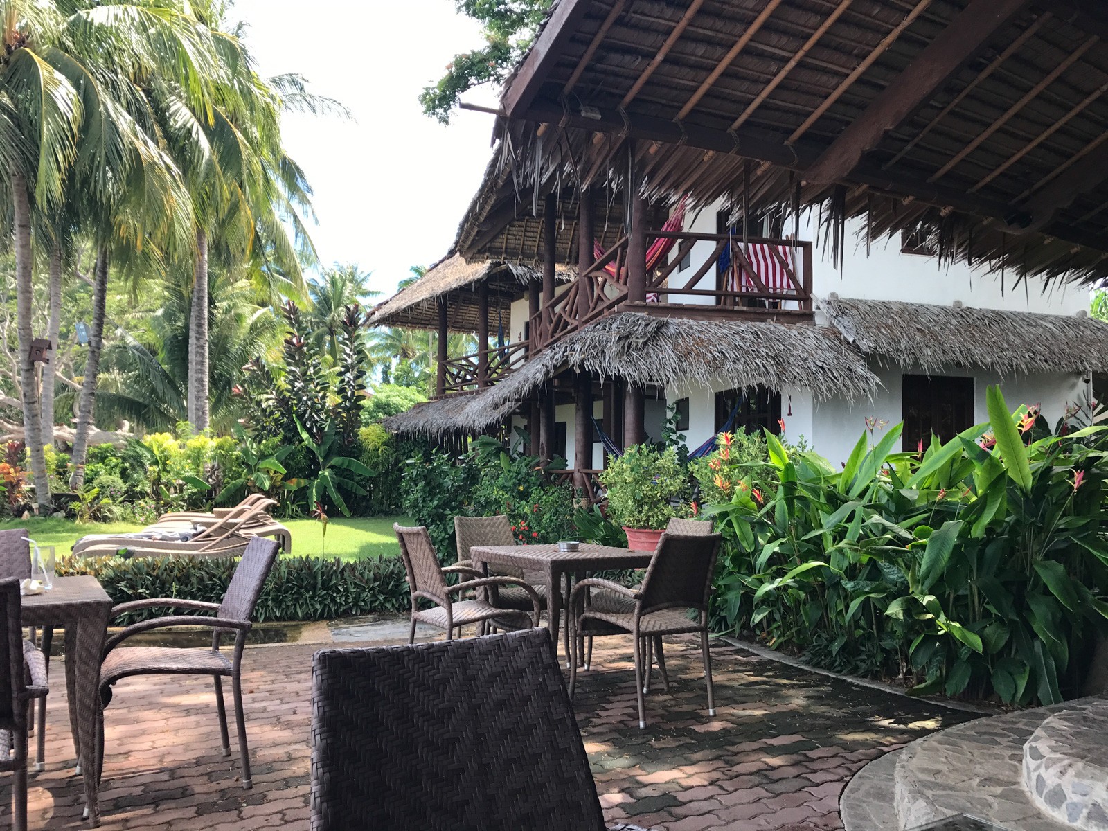 A tiled terrace with rattan chairs under a deep thatched eave, planting and palms crowding the edge