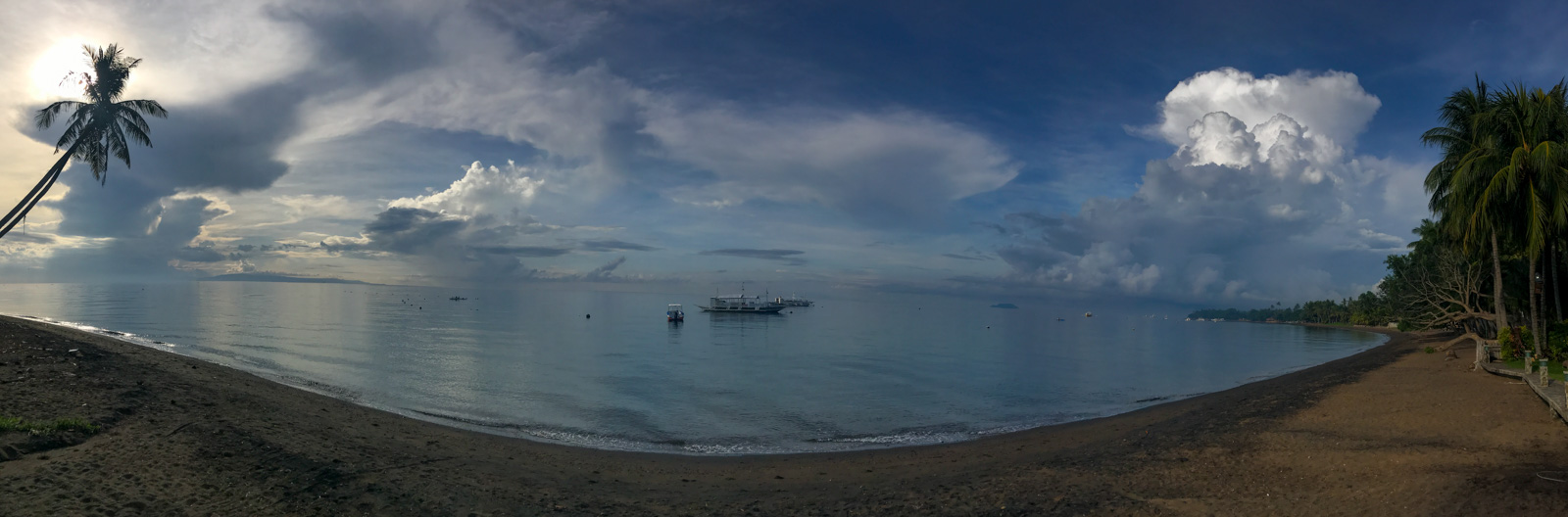 The black sand shore under building cloud, a boat anchored off and the palms leaning over the beach.