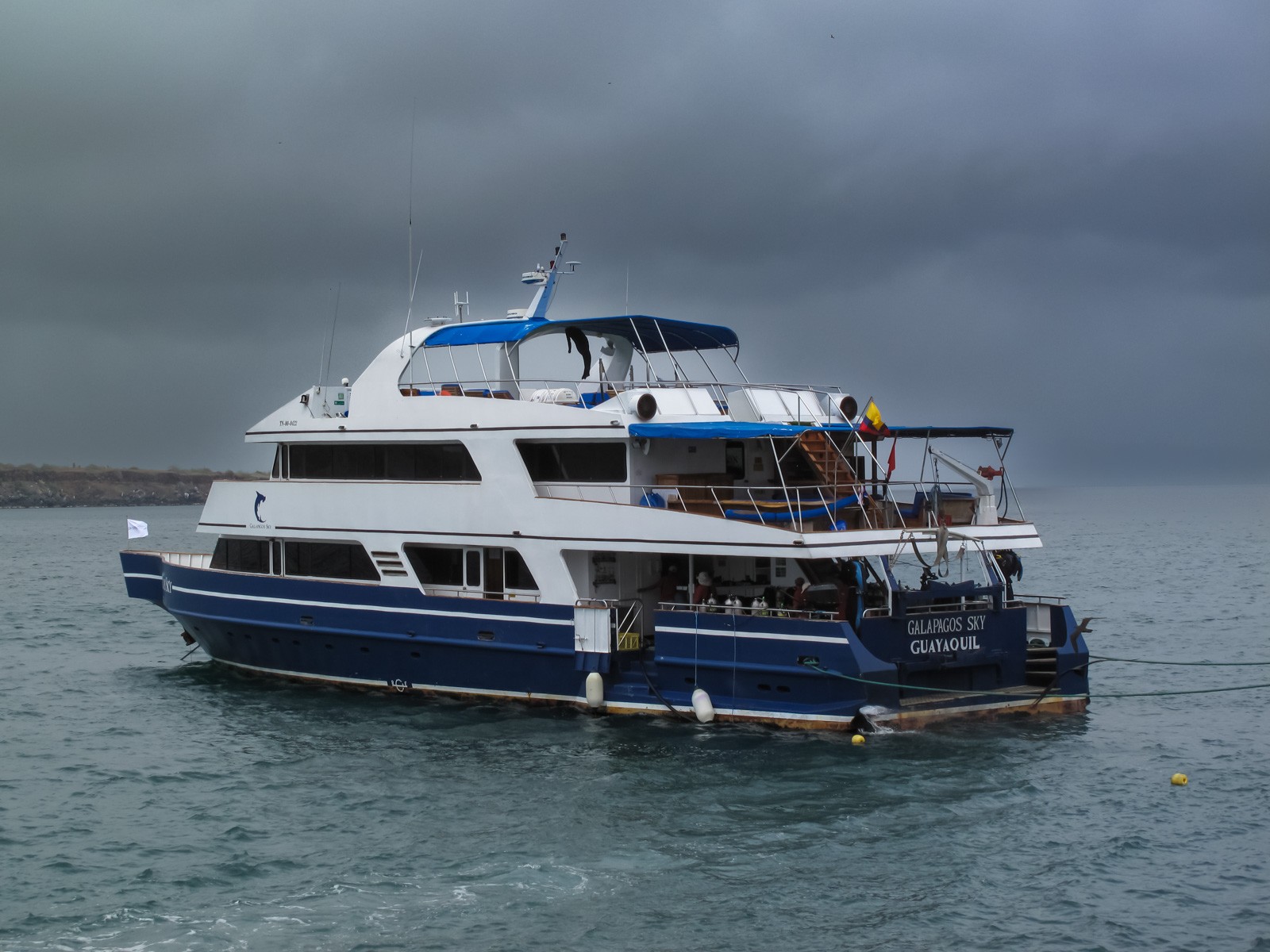A white liveaboard with a blue hull stripe lying at anchor under heavy grey cloud