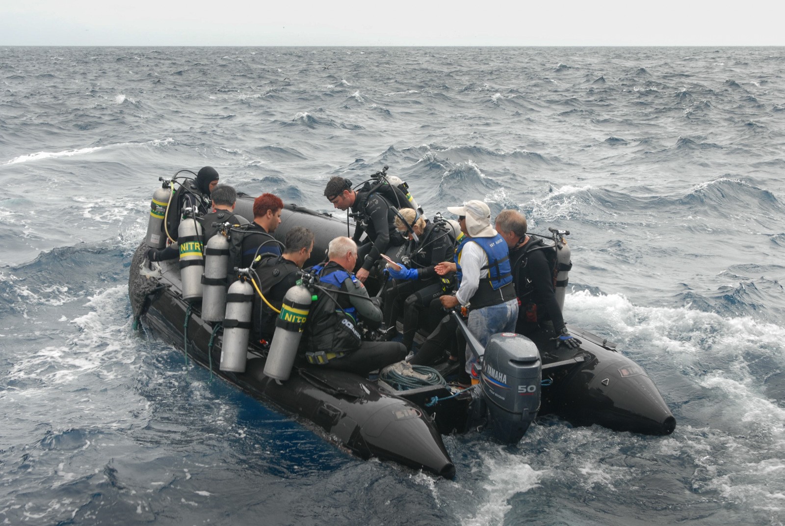 Divers in wetsuits packed shoulder to shoulder in an inflatable tender, tanks upright between them, running through open swell