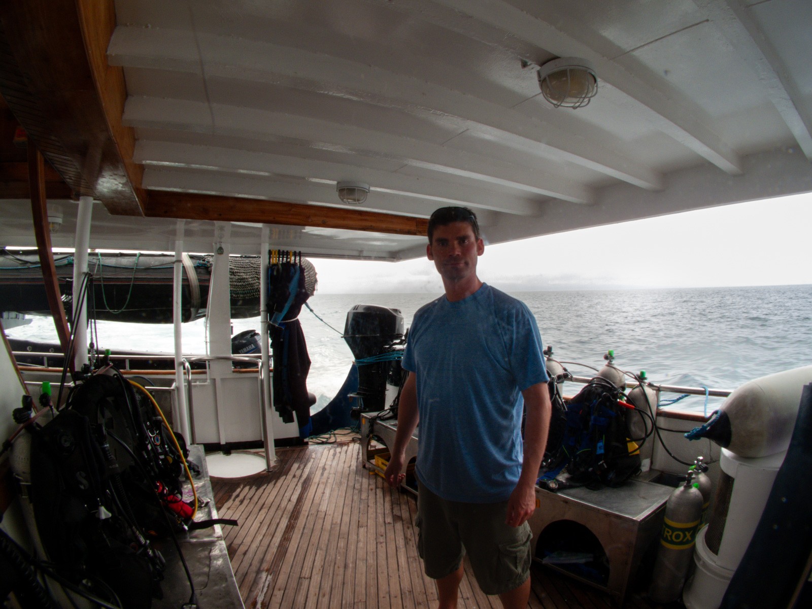A man standing on the covered dive deck of a liveaboard, gear stowed along the rail behind him and the sea running past