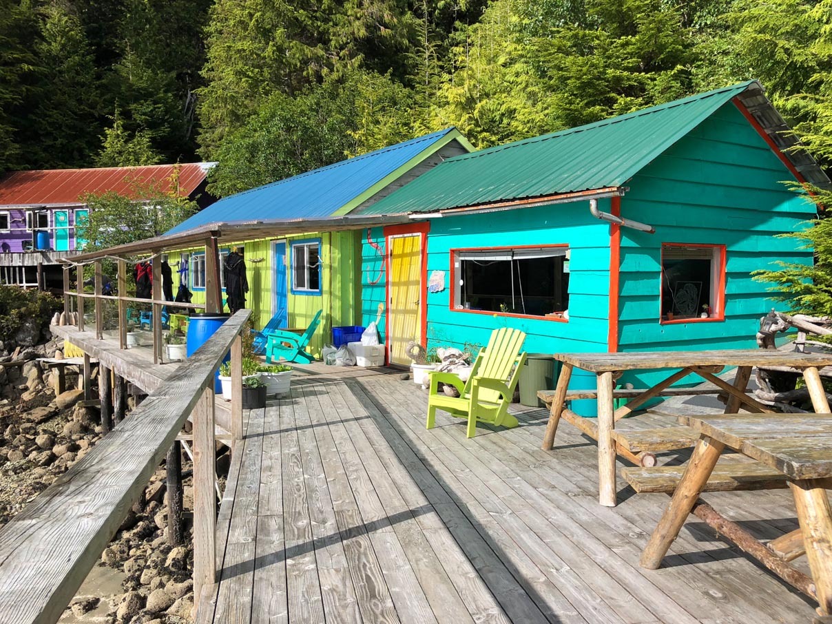 A timber boardwalk running past brightly painted cabins in teal, red and green, with deck chairs and a trestle table out front