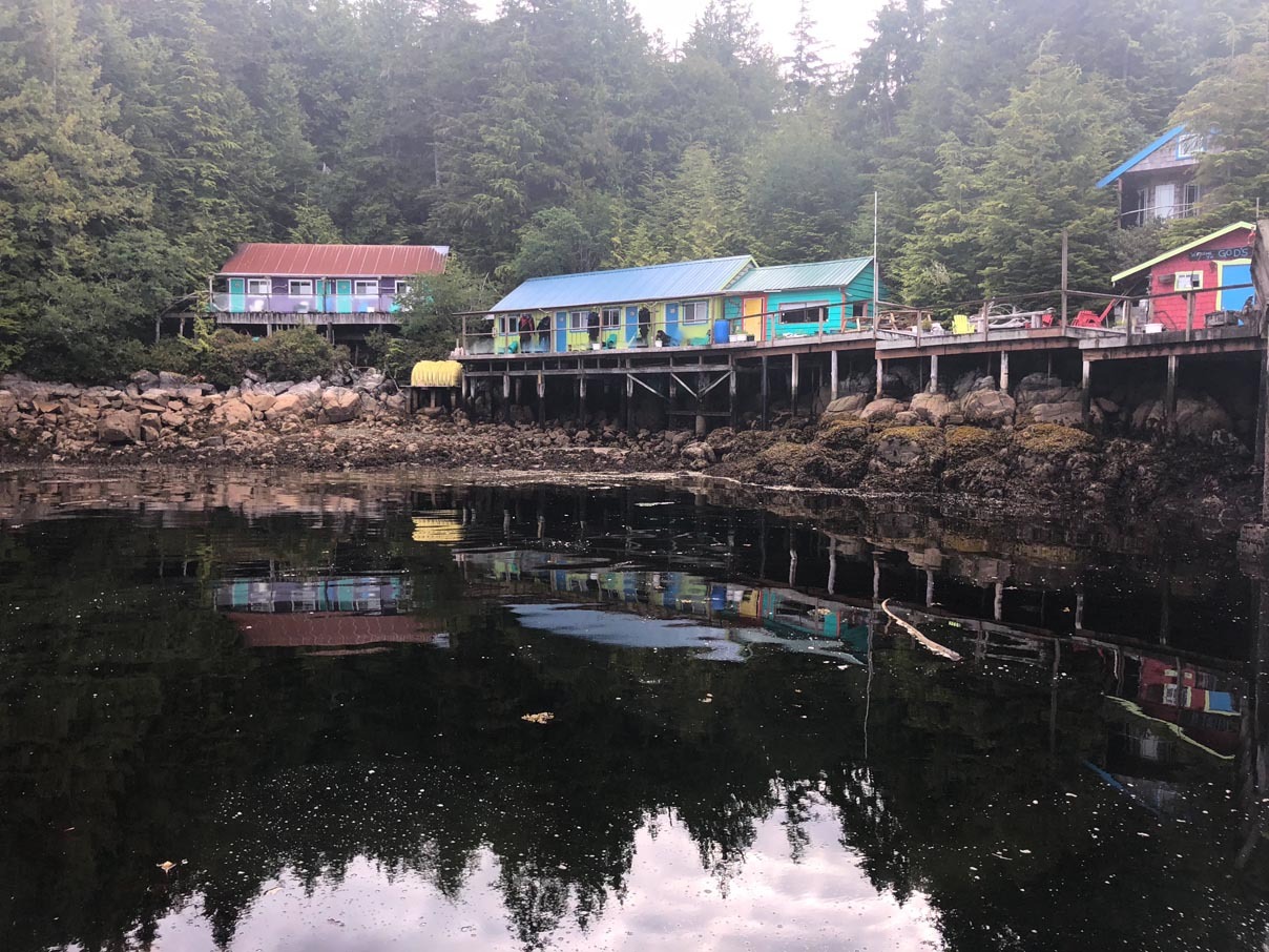 The whole row of painted lodge buildings seen from the water, standing on pilings under dense coastal forest