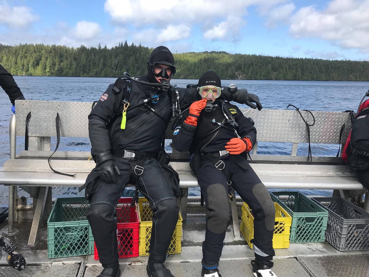 Two divers in drysuits and hoods sitting on the bench of a dive boat, milk crates of gear stacked beside them