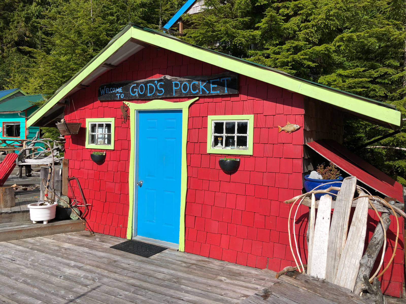 A red shingled hut on the dock with a bright blue door and a hand-painted welcome board over it, hose and gear stacked along the wall.