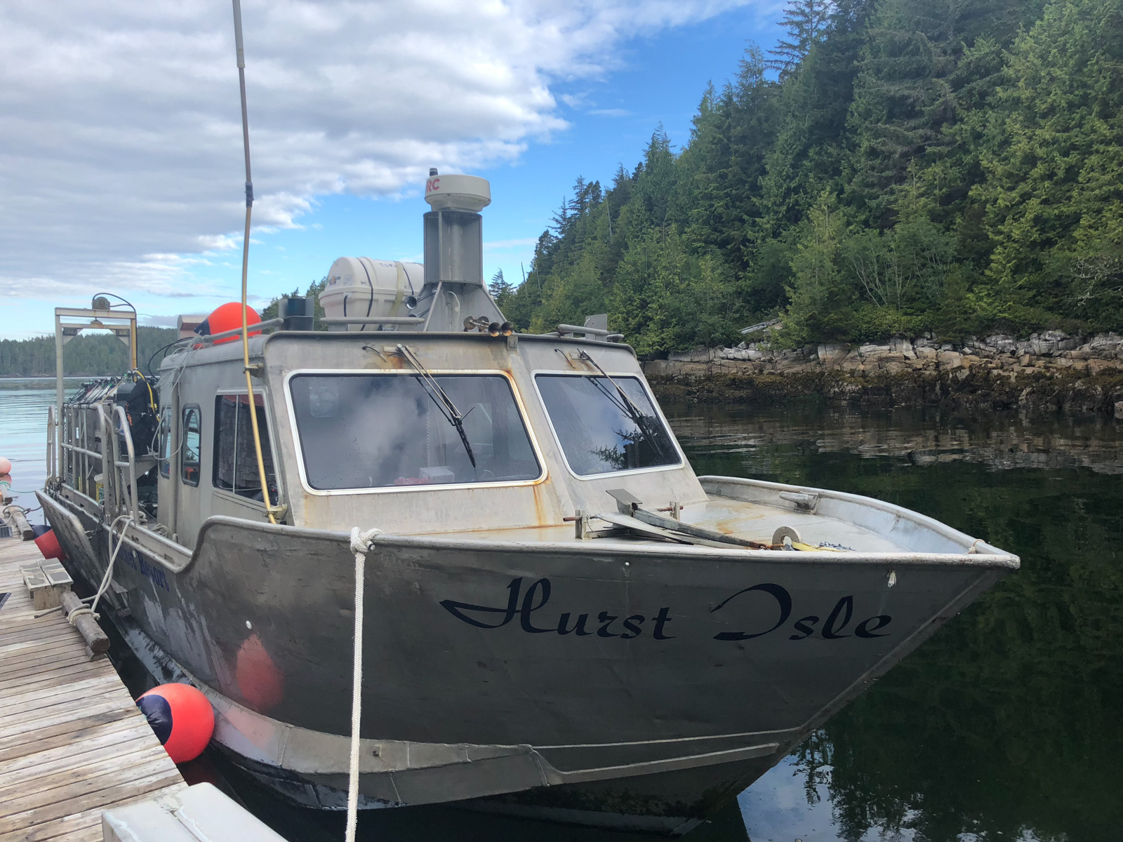 An aluminium dive boat tied alongside, her name lettered on the bow and the treeline running down to the water behind.