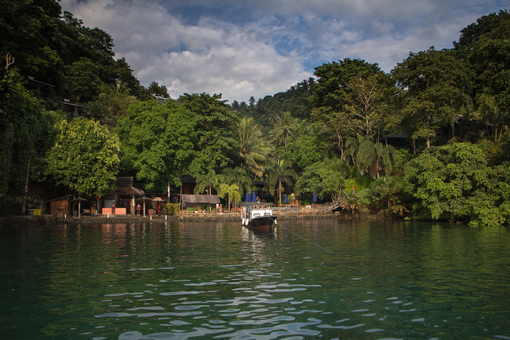 The resort seen from the water, low buildings set into steep green hillside with a boat moored in front