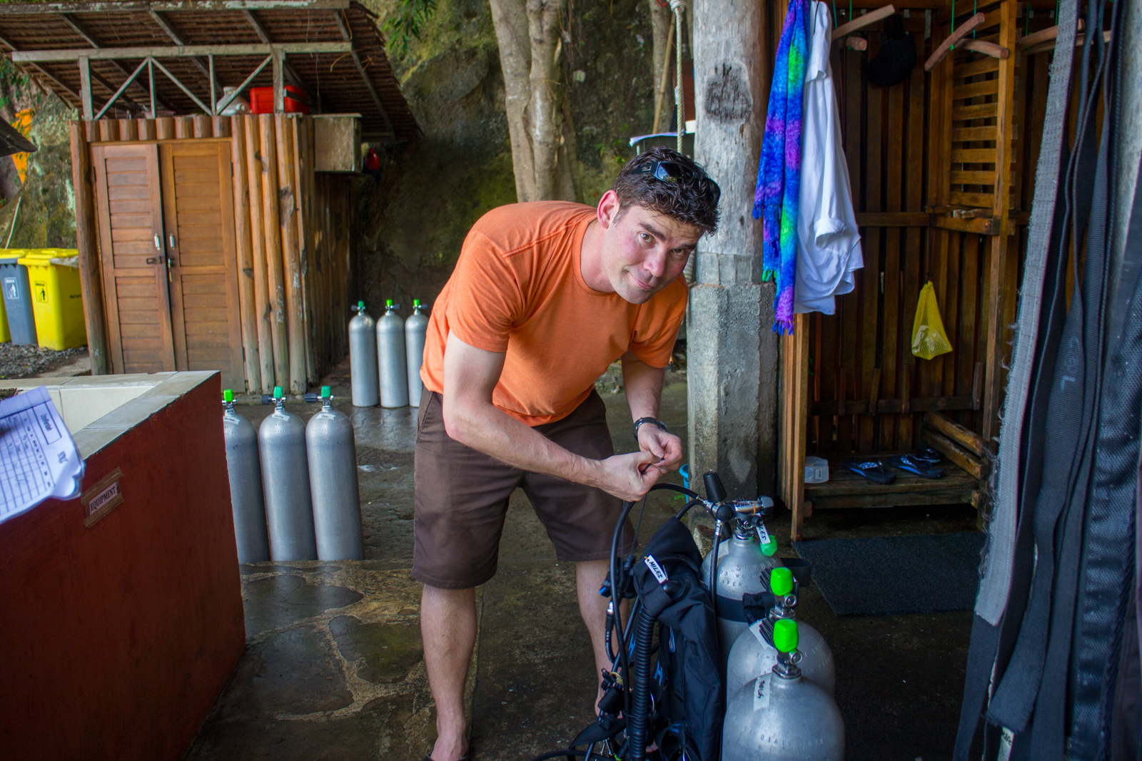 A diver clipping a regulator onto a cylinder in the tank room, rows of filled tanks standing against the back wall.