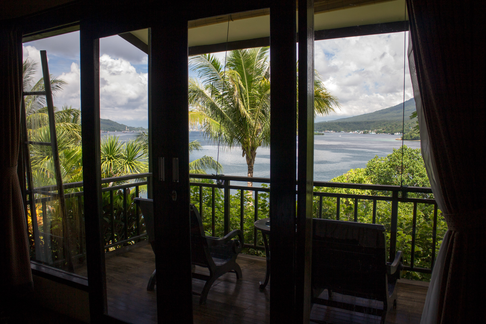 Sliding doors open from a room onto a balcony, the strait and the far volcano filling the gap between the palms.