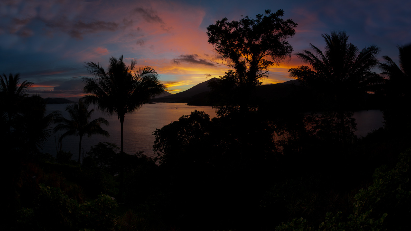 Sunset across the strait, the volcano and the palms gone to silhouette against an orange sky.