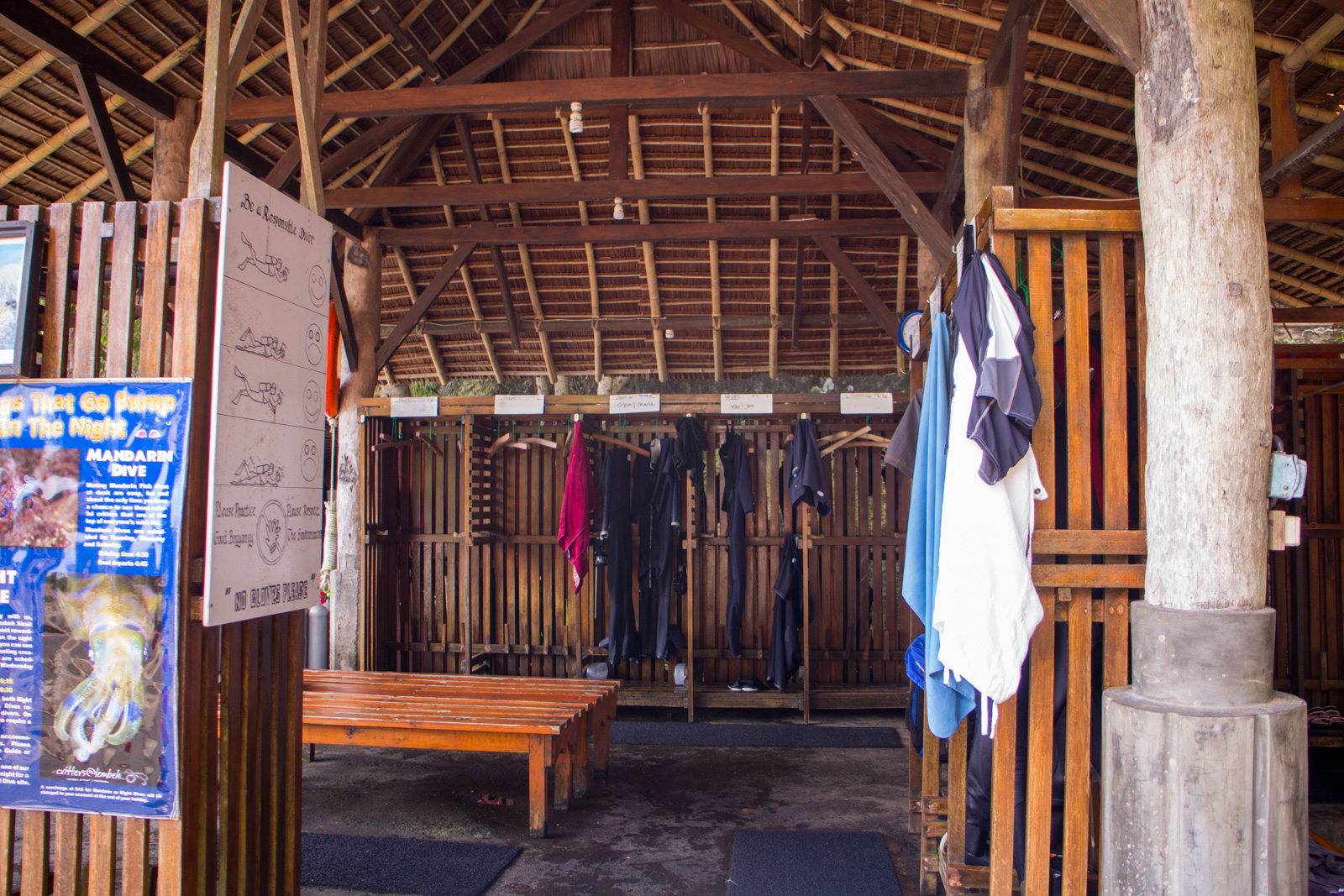 An open-sided timber drying room, wetsuits and towels on hangers along the rail, a bench down one side and dive notices pinned by the door.