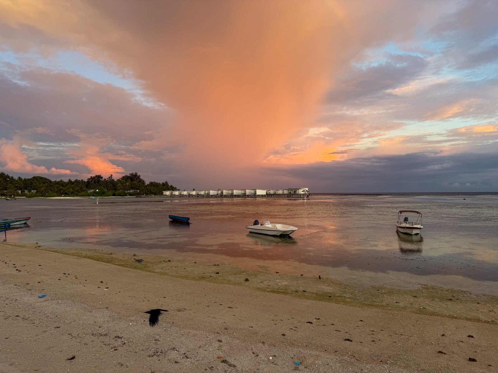 Sunset over the lagoon at low water, small boats moored off the sand and a line of overwater rooms in the distance.