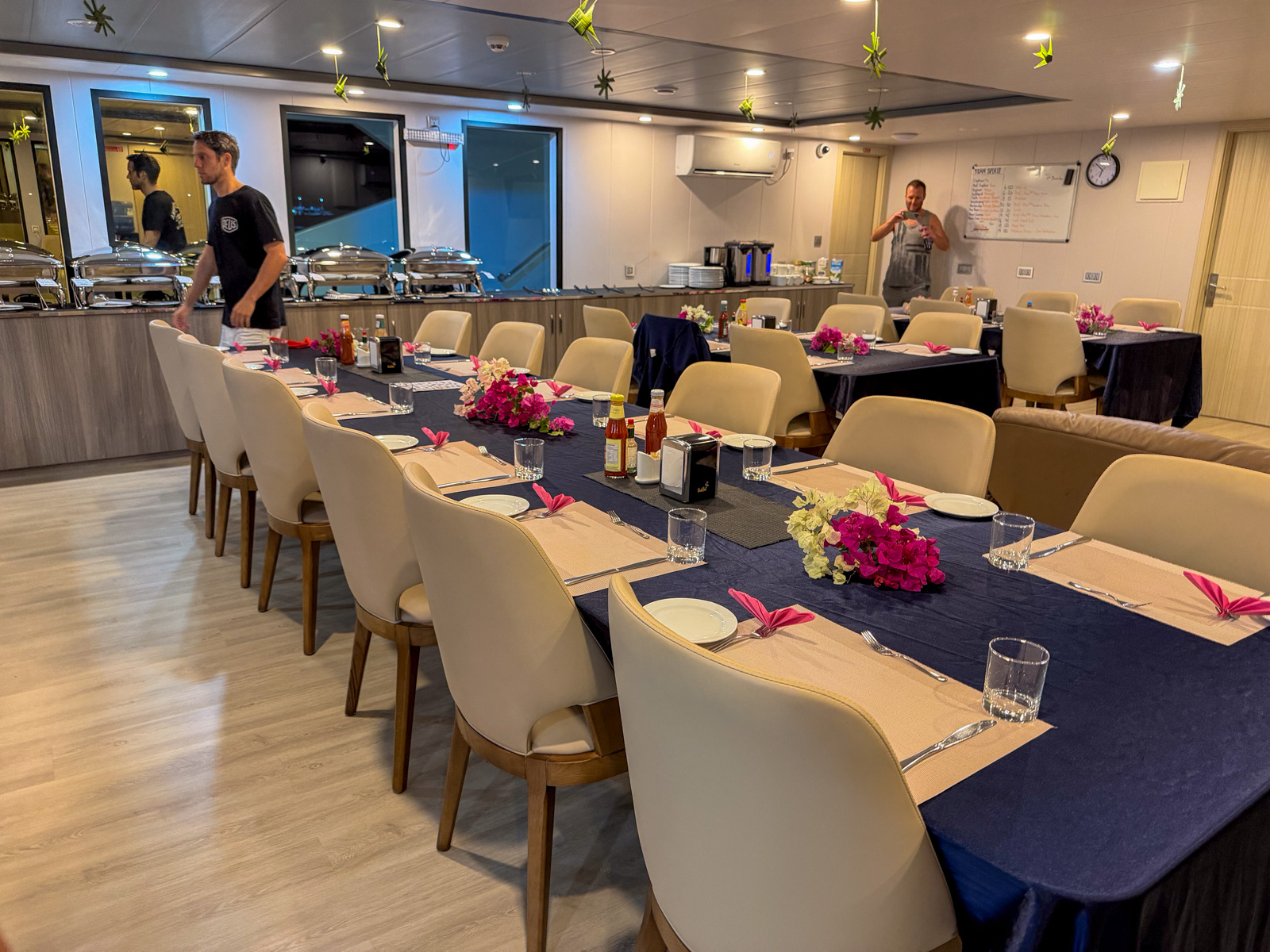 The saloon laid for dinner, one long table down the middle with flowers and wine set out and crew working the buffet behind.
