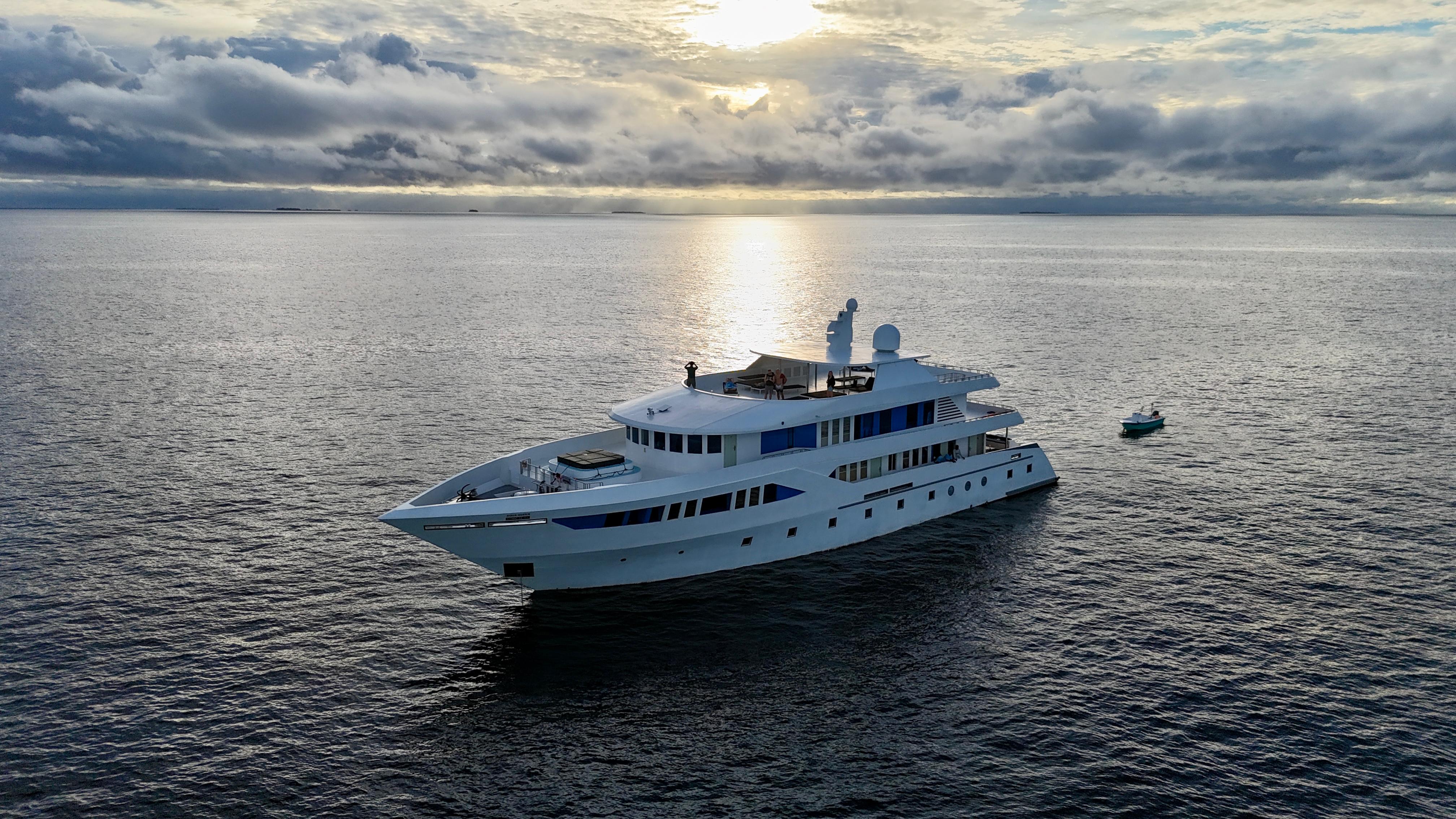 The boat lying at anchor in flat water under a heavy dawn sky, her tender alongside.