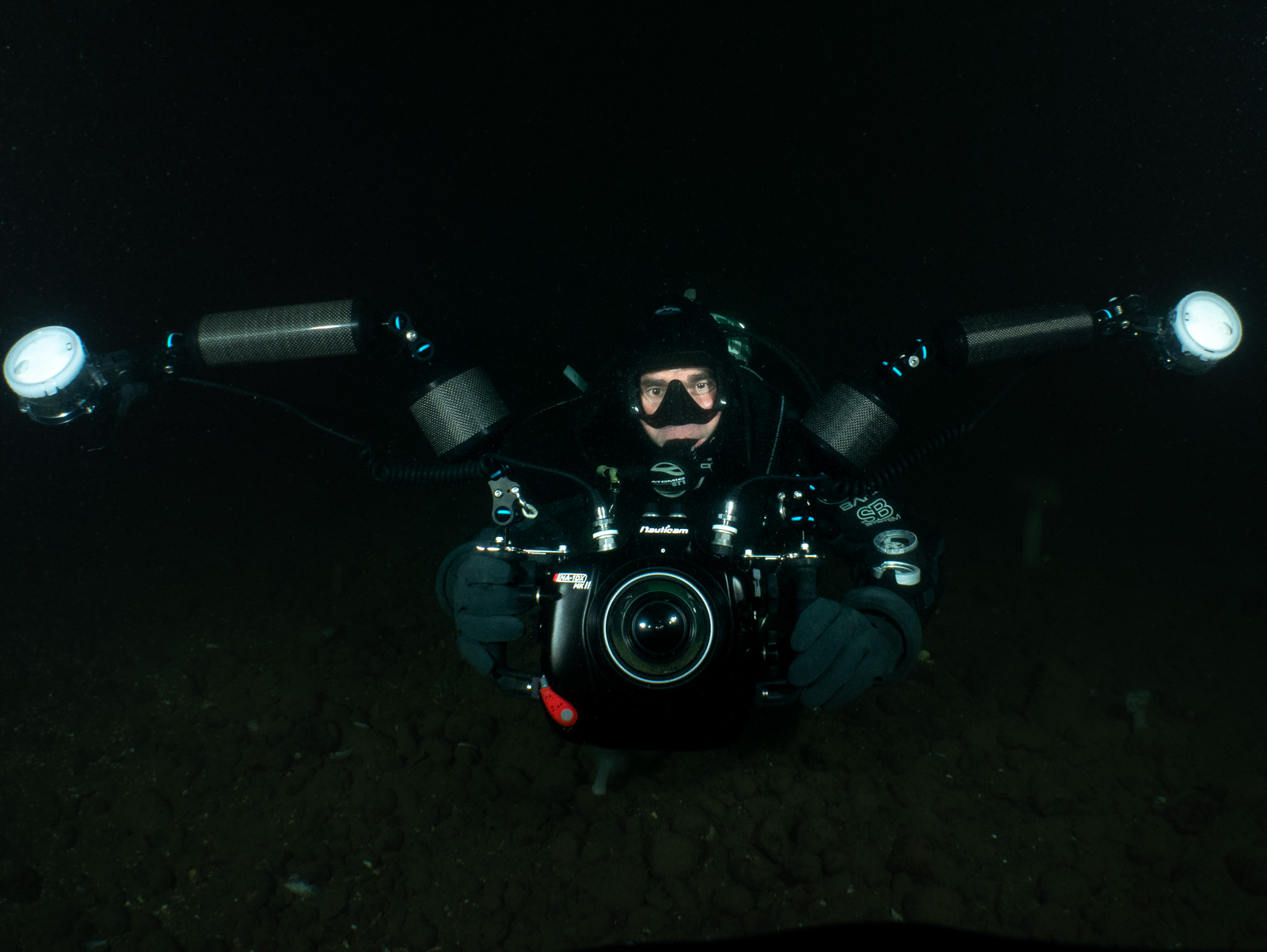 Miles in a drysuit and hood on a black night dive, both strobes turned toward the camera over a dark cobble bottom