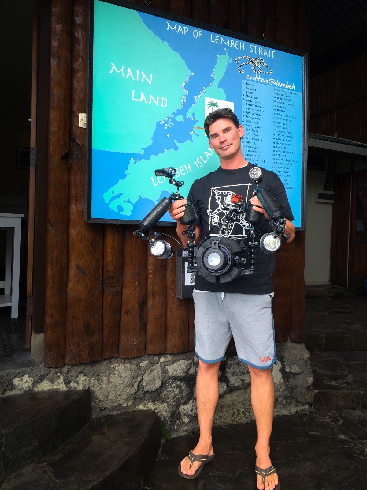 Miles on the dock under a hand-painted map of Lembeh Strait, holding his camera housing with two strobe arms and focus lights spread wide