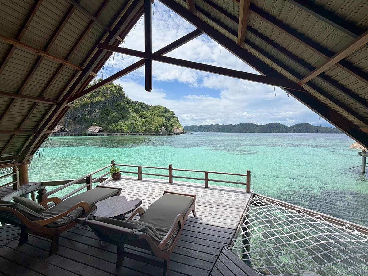 The view from inside an over-water bungalow, timber deck and loungers framed by the roof beams, a limestone island across the bay