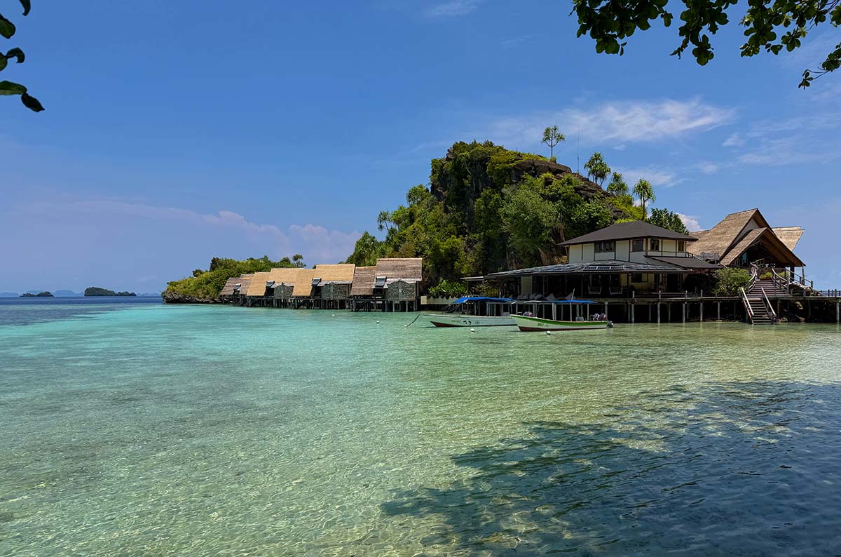 The resort from the water: a row of over-water bungalows on stilts along the shore, the main building on a limestone headland behind them, a dive boat moored in the turquoise shallows