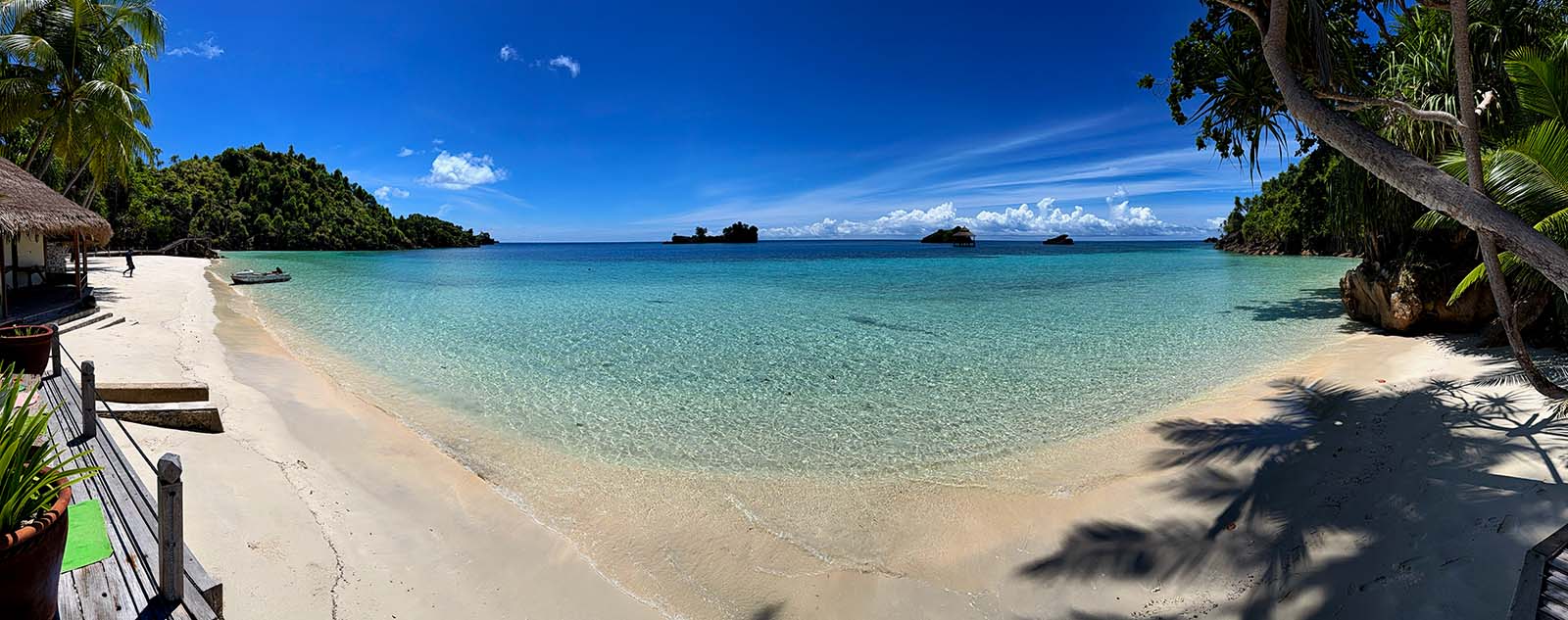 The resort's beach in a panorama: white sand curving round a turquoise bay, a dinghy pulled up at the water's edge, palms and the deck of a bungalow on the left, small limestone islands on the horizon