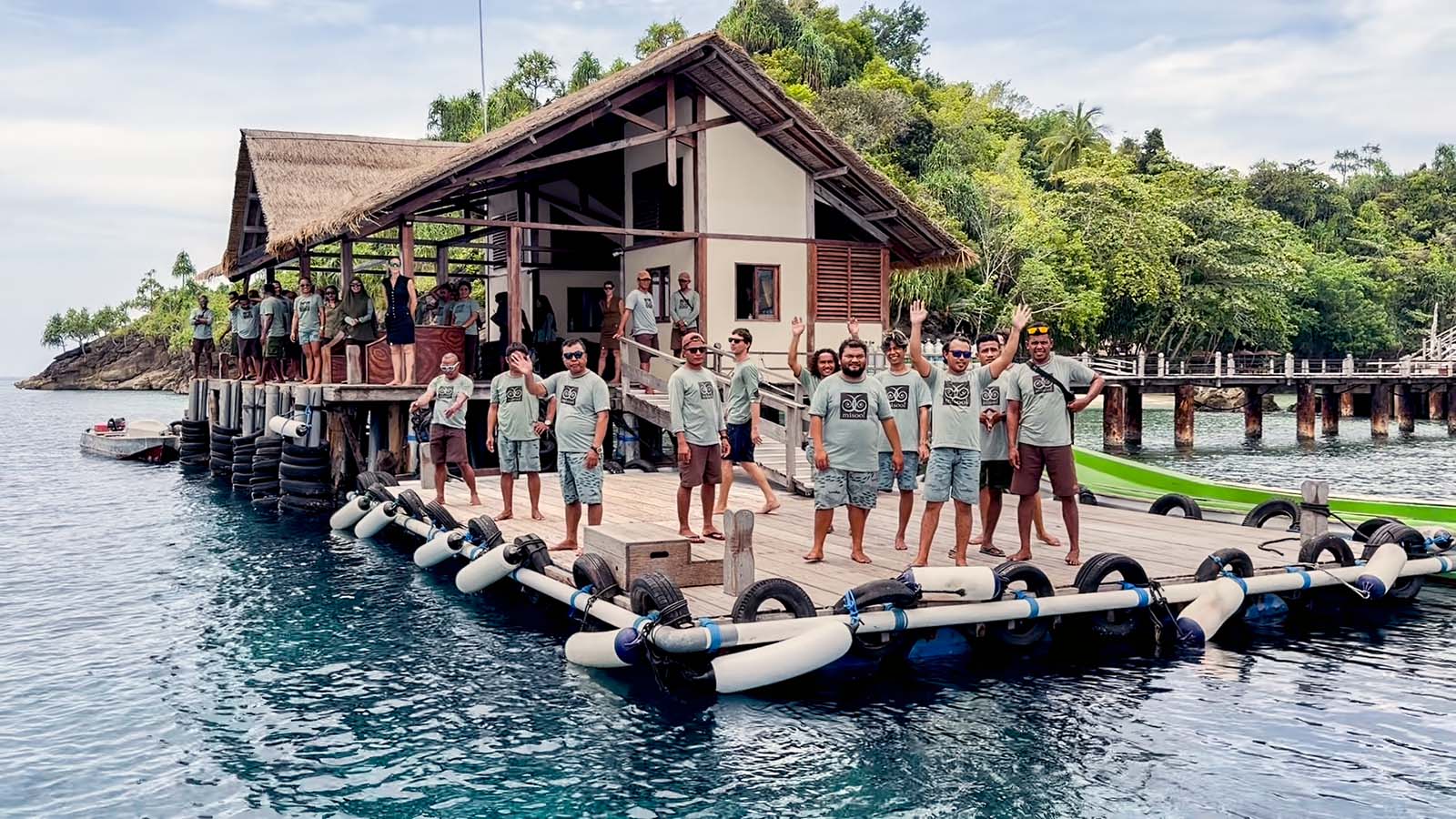 The Misool Resort team on the floating jetty, a dozen staff in matching resort shirts waving from the dock in front of the thatched dive centre, the boardwalk and forested headland behind