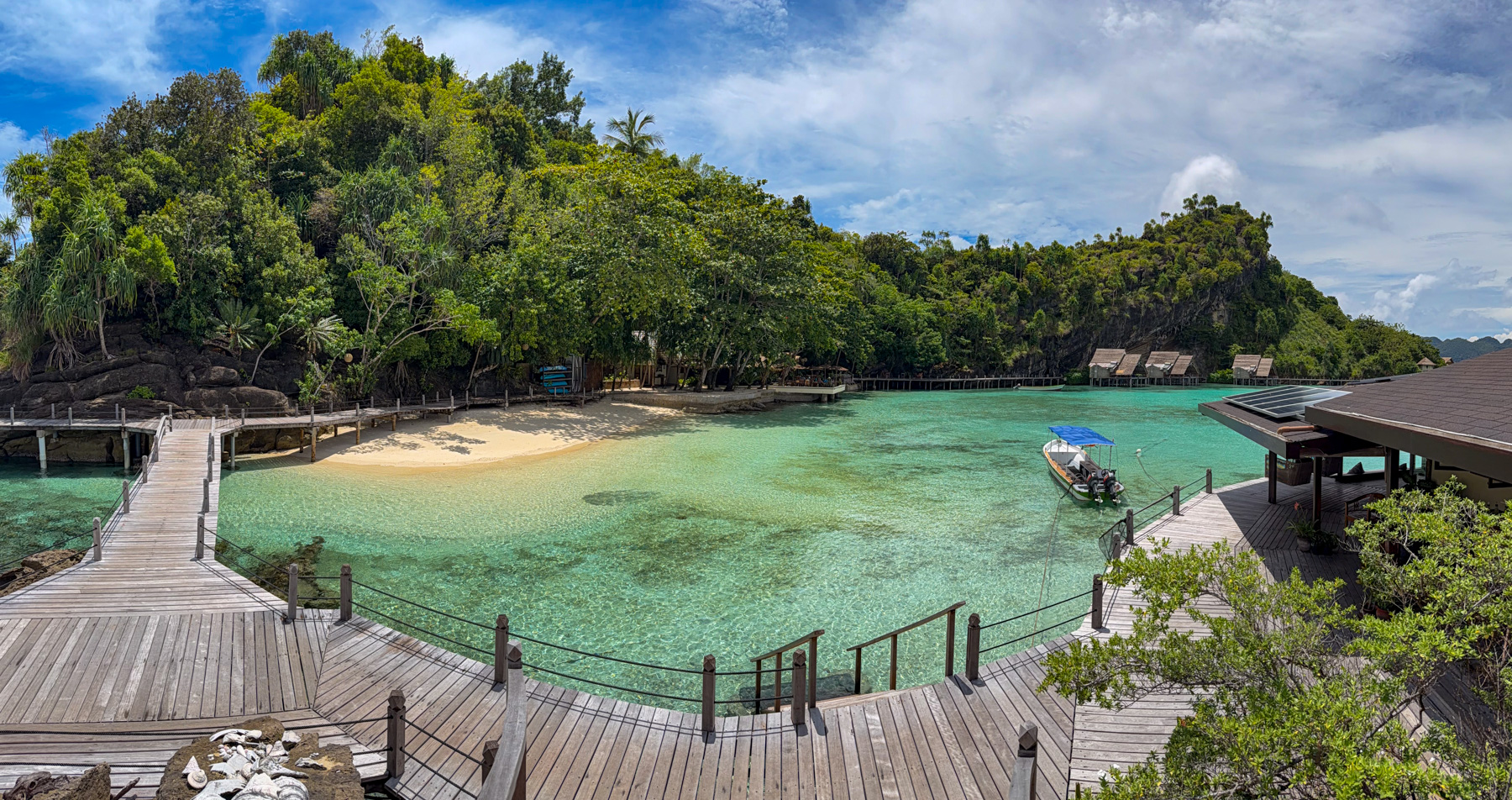 A boardwalk curving round a shallow bay in panorama, a strip of sand at its head, a boat under a blue canopy moored off an open timber building on stilts, thatched bungalows along the far shore
