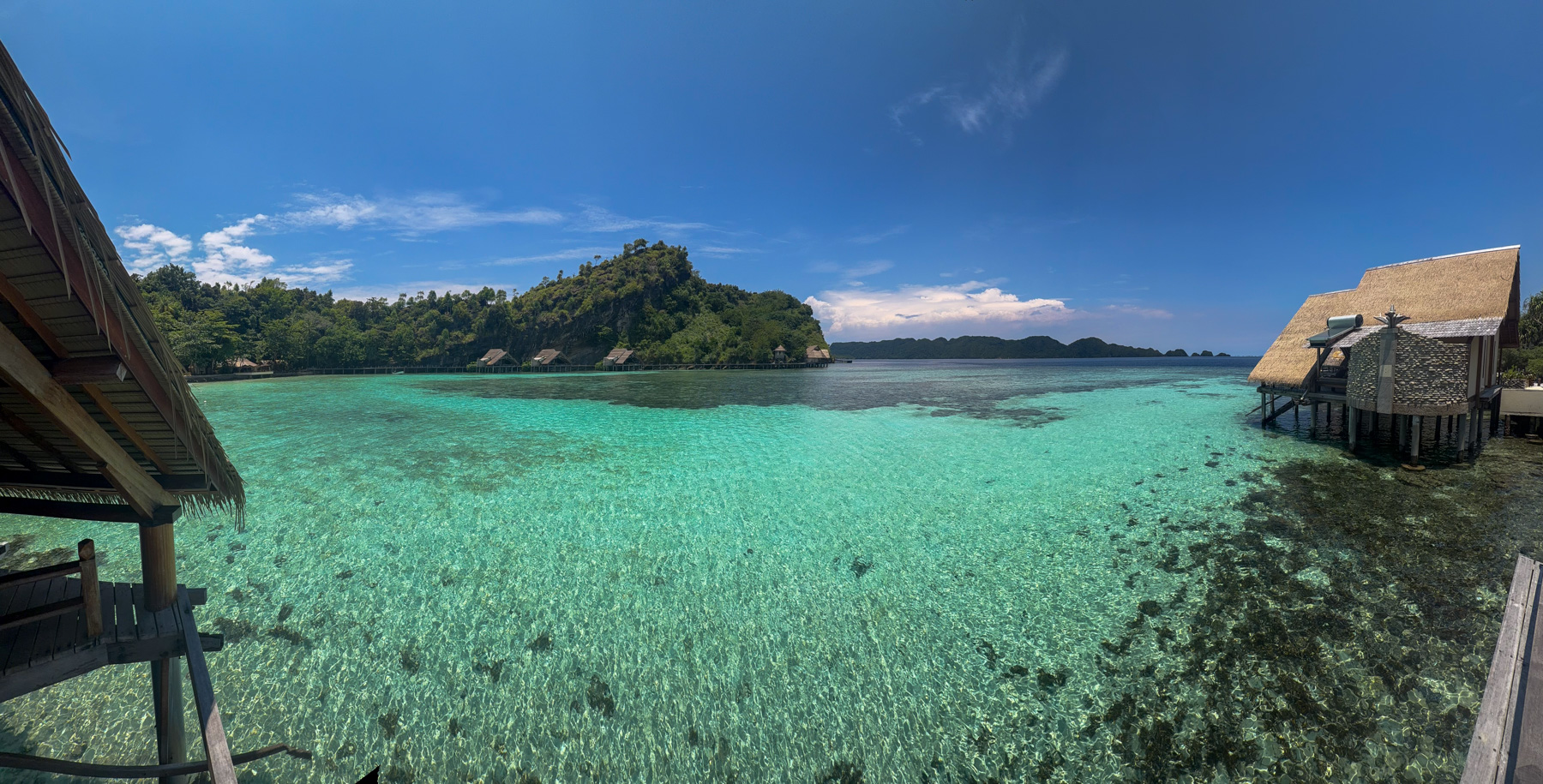 A panorama across the reef flat from under a thatched eave, coral heads showing through the shallows, a line of bungalows on stilts along the shore and one standing clear of the rest on the right