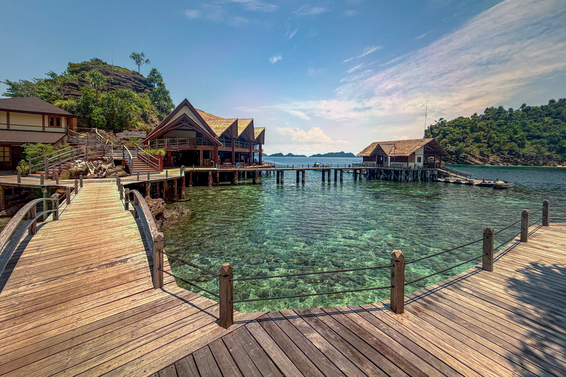 The main boardwalk running in to a tall thatched building with a rope-railed deck, a smaller hut out at the end of the jetty and boats tied alongside, coral showing under the shallows