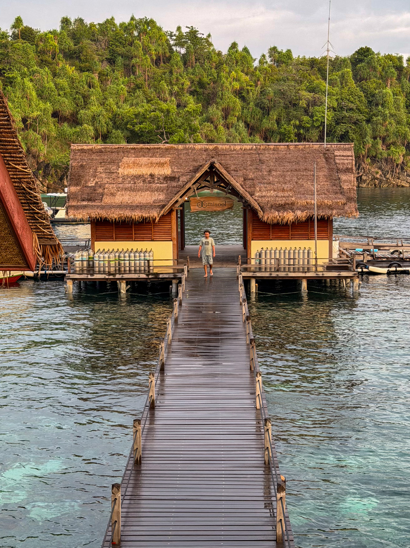A jetty running out to the thatched dive centre in evening light, ranks of scuba cylinders standing along both open sides and a man walking in under the gable