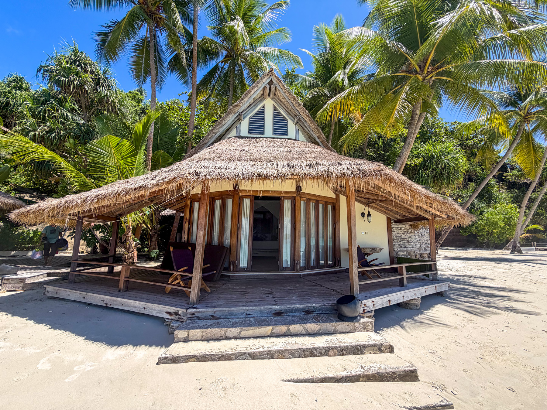 A beach bungalow with a steep thatched roof and folding doors open onto its deck, a chair at each end and stone steps down to the sand, coconut palms behind