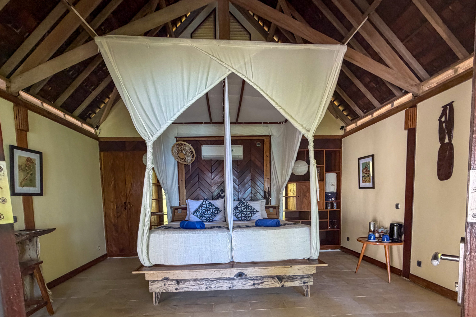 The inside of a bungalow: a bed under a white mosquito net, an open timber roof above it, a chevron-panelled headboard wall and a tray of tea things on a side table